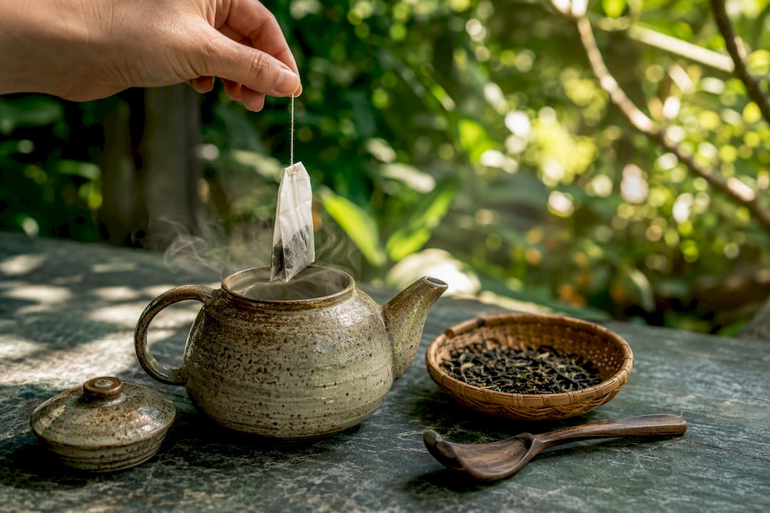 Hand placing tea bag into teapot with loose leaf tea nearby