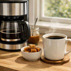 Caramel flavored coffee in white mug with coffee maker and caramel cubes on wooden table near window