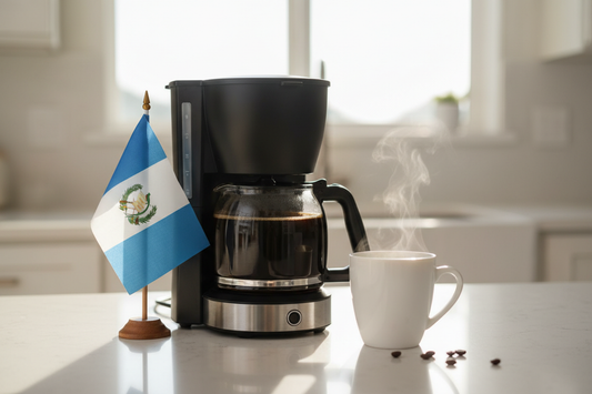 lifestyle picture with a coffee maker on a kitchen counter with a half-filled coffee pot with the Guatemala flag on one side and a cup of brewed coffee on the other side